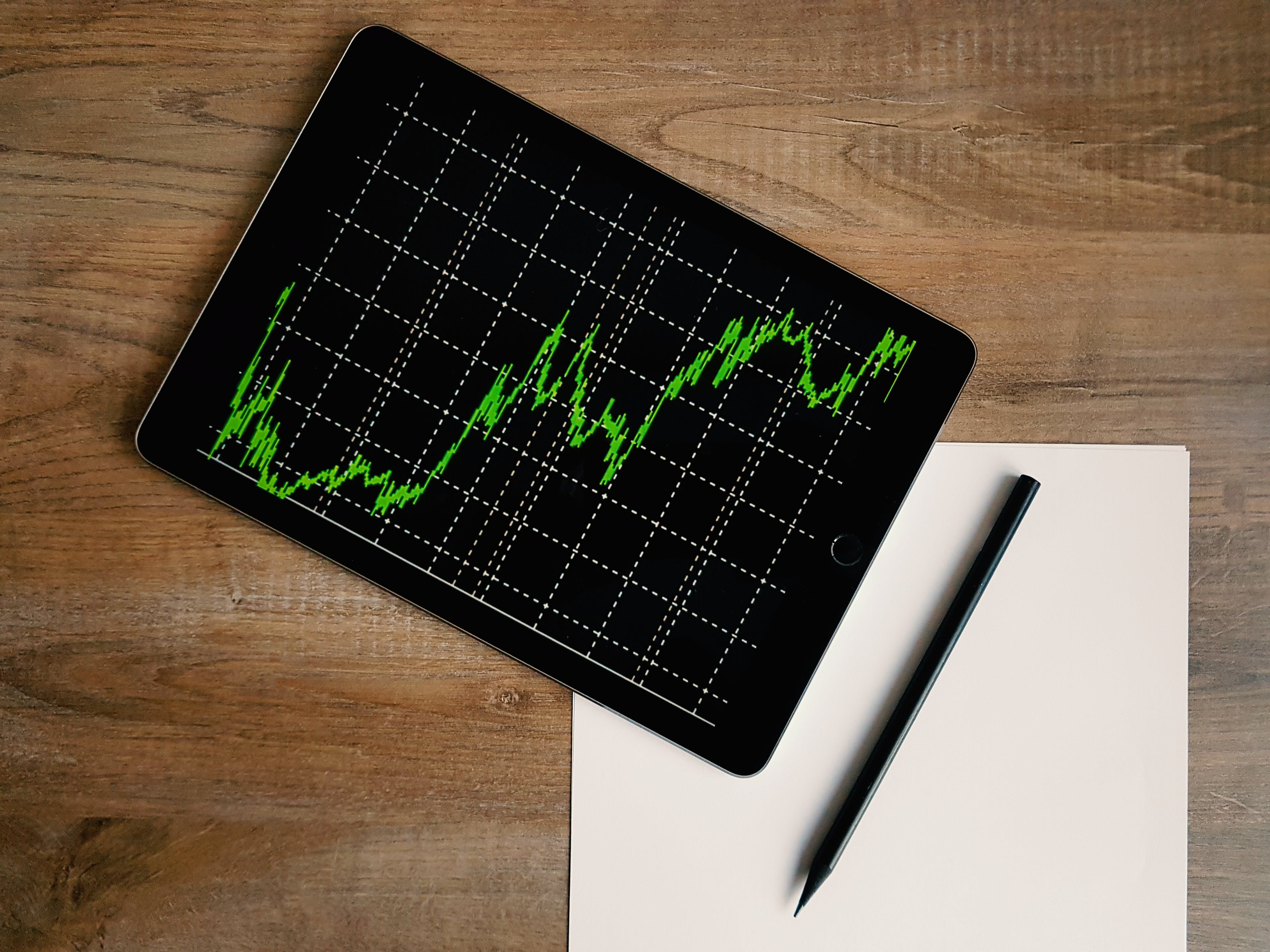 a modern tablet with a green stock market graph displayed on a black background. The graph is represented by bright green lines against a grid. The tablet is sleek and modern, with no visible buttons indicating it might be operated by touch. A stylus pen lies parallel to the right edge of the tablet; it’s dark-colored, slim and appears to be designed for precision. Underneath the stylus, there’s an unmarked sheet of white paper that hasn’t been written or drawn upon yet. All items are placed on a smooth wooden surface that has natural light brown tones and wood grains visible.