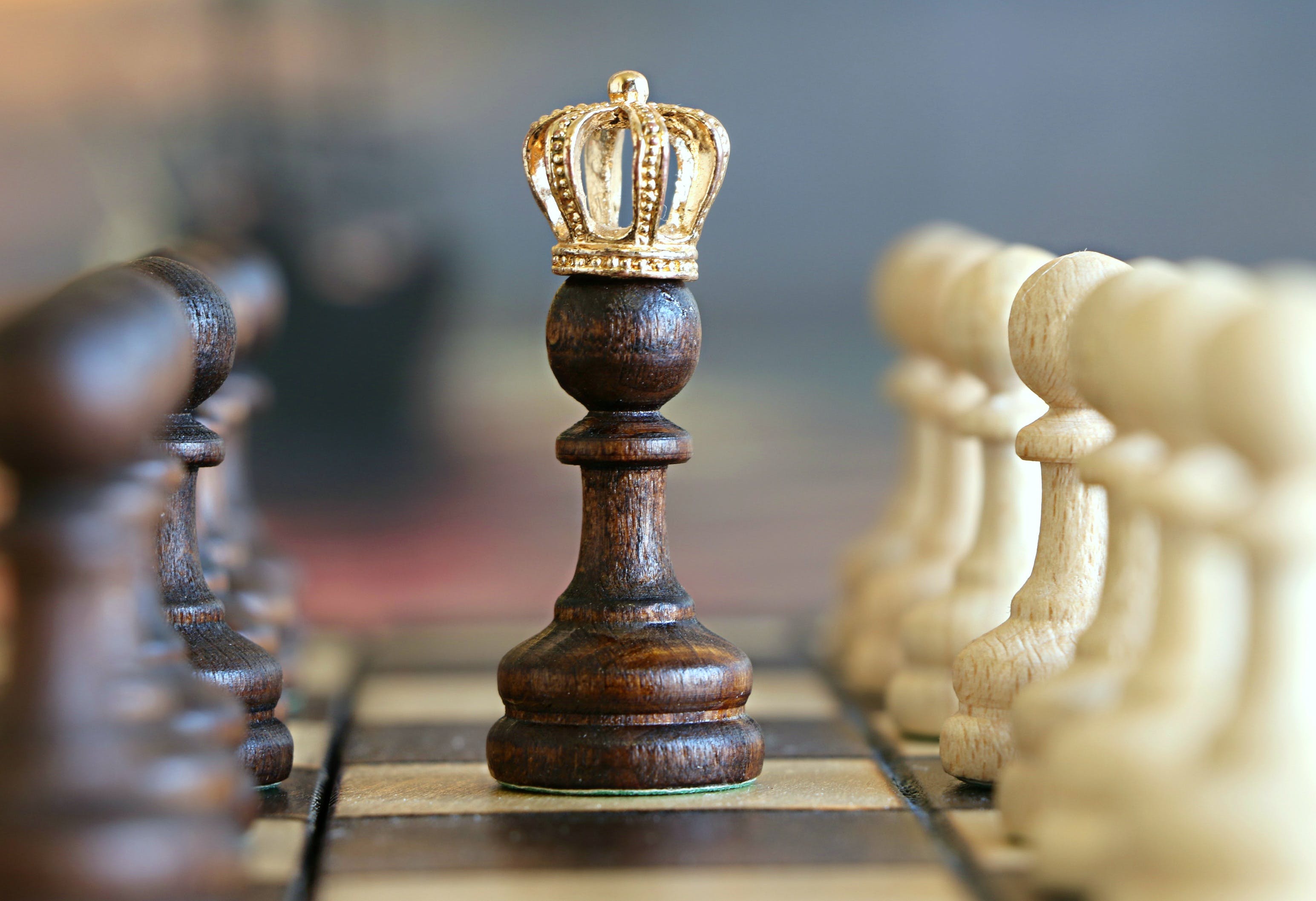 a dark wooden pawn on a chessboard. The pawn is adorned with a golden crown and is positioned between rows of black and white chess pieces, creating a stark contrast in colors. The intricate details of the crown and the wood grain on the pawn are visible, highlighting the quality of craftsmanship. The background is blurred and indistinct, drawing attention to the crowned pawn in sharp focus at the center. There’s an evident contrast between the dark and light-colored chess pieces as well as between them and their environment.